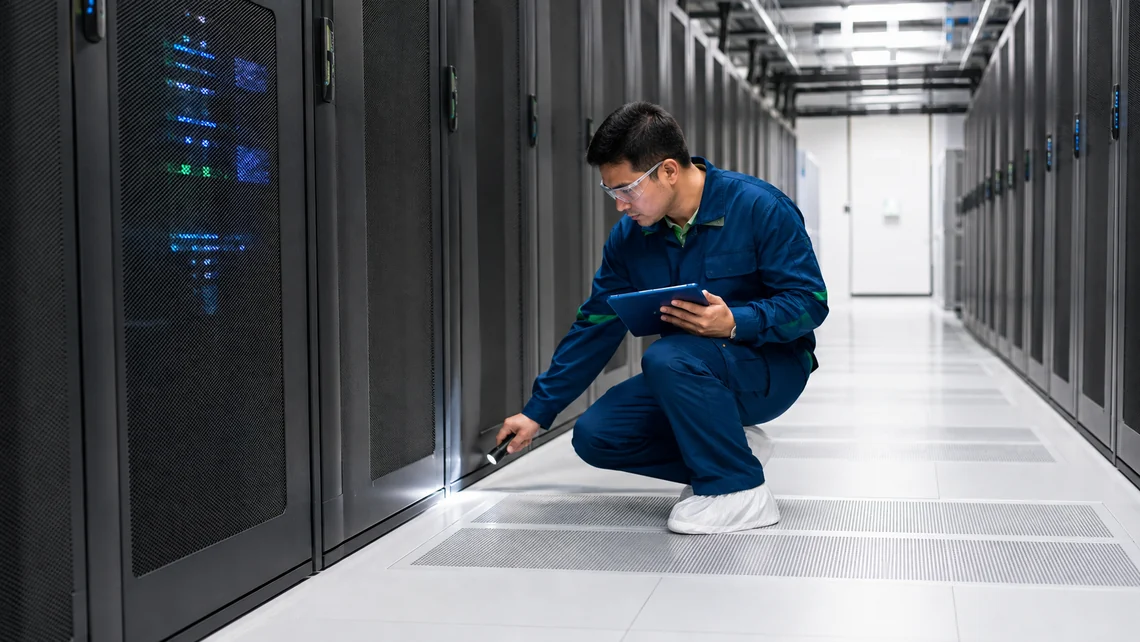 Illustration. A technician crouches beside a row of dark server cabinets holding a tablet in one hand and shining a flashlight along the base of a cabinet door at a shallow angle, so that the beam rakes across the surface.