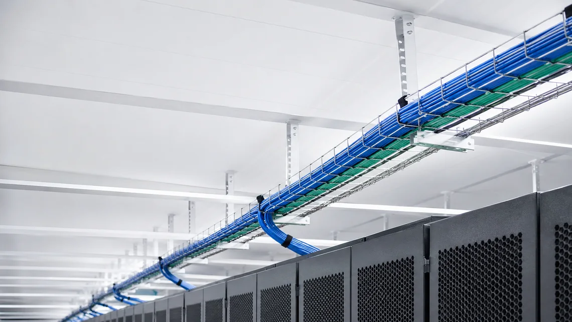Illustration. Wire-basket cable tray suspended from a data hall ceiling on threaded rod and strut, carrying bundled blue and green cabling above a row of dark server cabinets.